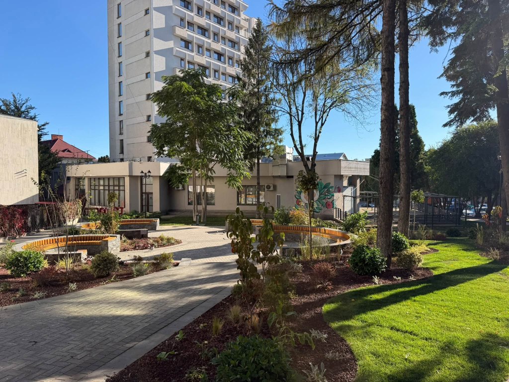 Peisaj urban cu o zonă verde amenajată, bănci din lemn și flori, în fața unei clădiri înalte.
