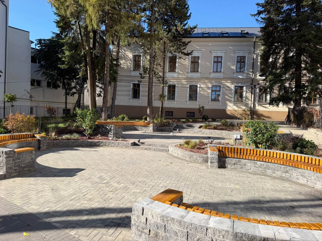 O zona de relaxare cu bănci din lemn și piatră, înconjurată de copaci, plante și o clădire istorică în fundal.