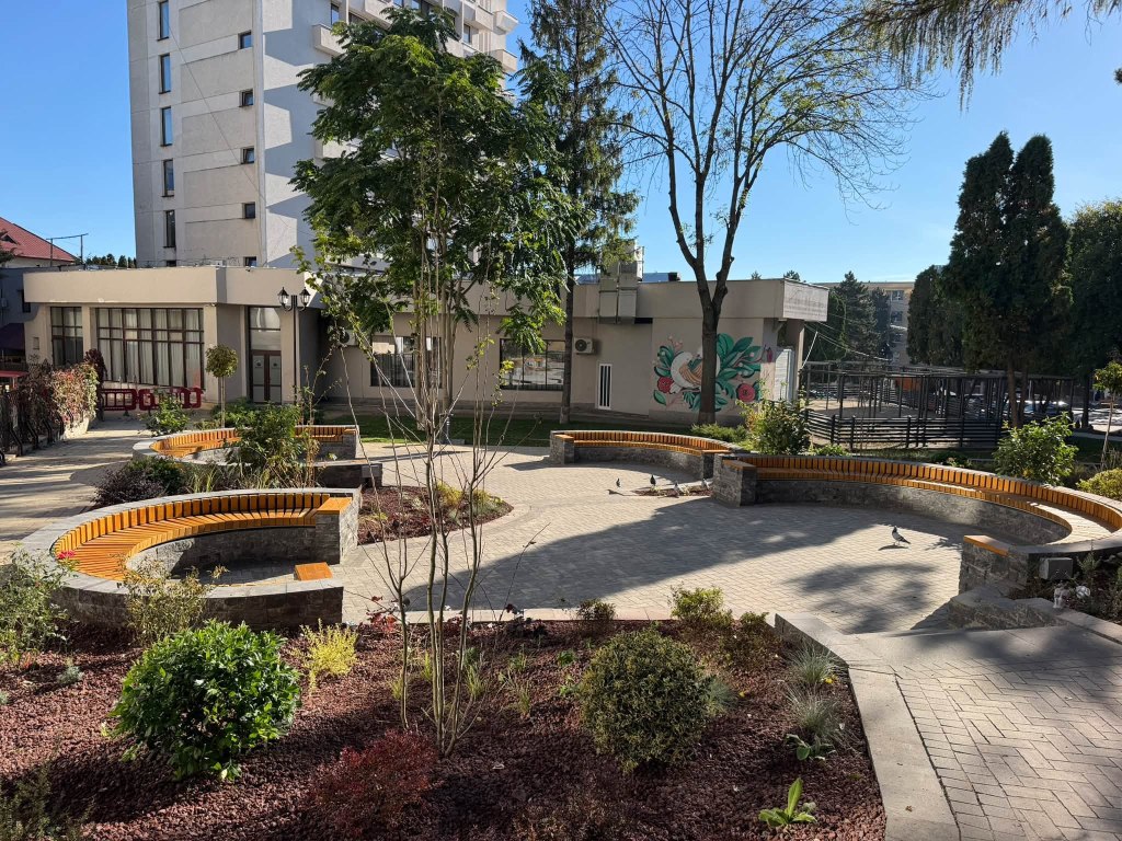 Un parc cu bănci rotunde din piatră, plante și arbori, situat lângă un clădire înaltă. Cer senin și păsări pe alee.