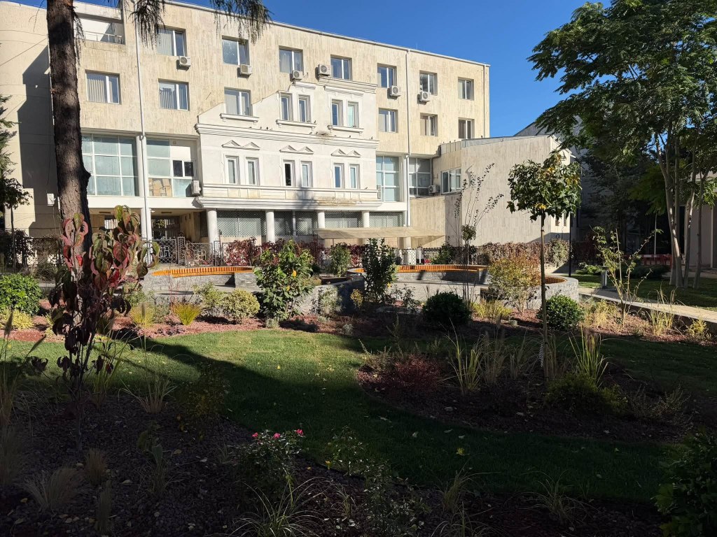Clădire modernă cu fațadă deschisă, amplasată într-un peisaj verde, cu o grădină amenajată și un mic bazin de apă, sub cerul senin.