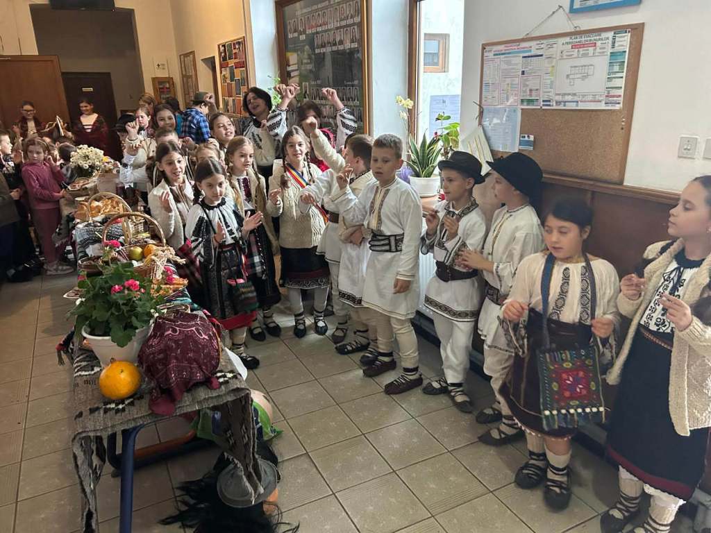 Grup de copii îmbrăcați în costume populare românești, participând la un eveniment cultural. Pe o masă sunt expuse diverse produse tradiționale, iar unii copii cântă și se bucură.