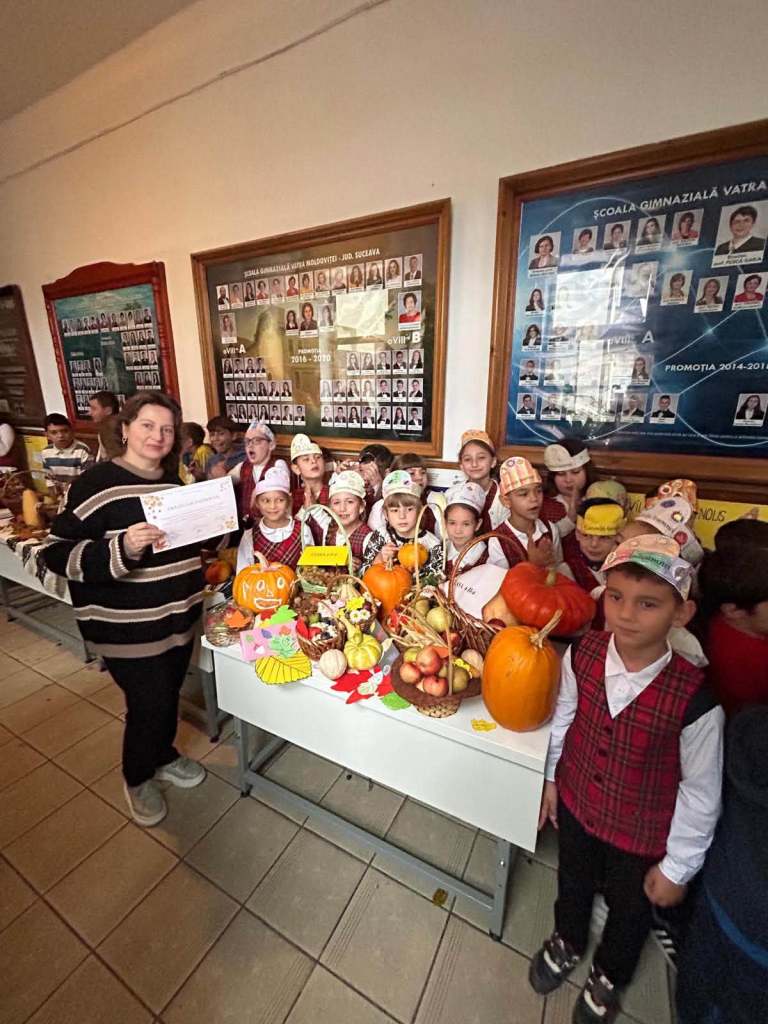 Un grup de elevi îmbrăcați în uniforme cu pălării colorate stau împreună cu un adult în fața unei mese pline cu fructe și legume, inclusiv dovleci. Pe perete, se află afișe cu fotografii ale elevilor.