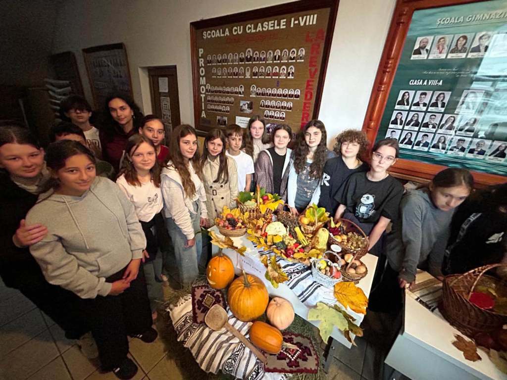 Grup de elevi și profesori pozând în fața unui set de mese decorate cu fructe și legume, într-o școală.