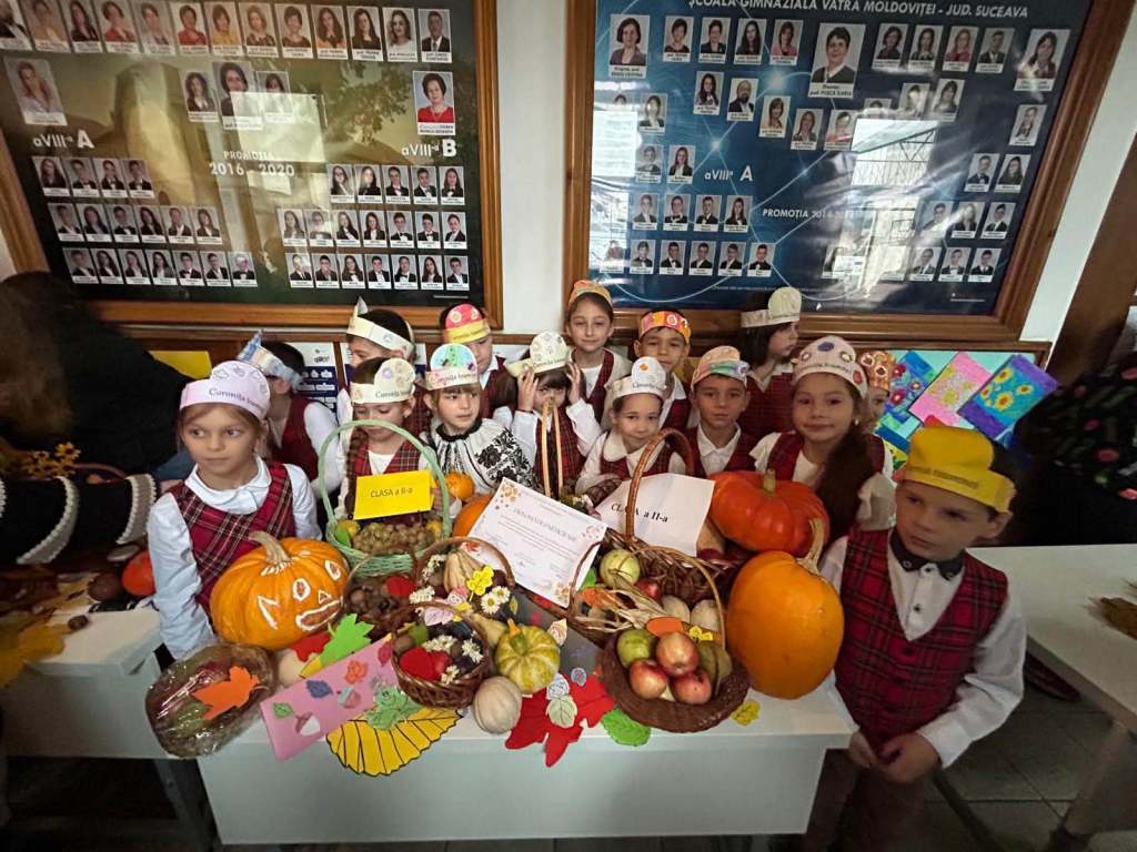 Grup de copii îmbrăcați în costume tradiționale, pozând lângă un stand cu dovleci, fructe și decorațiuni de toamnă, într-o sală de clasă.