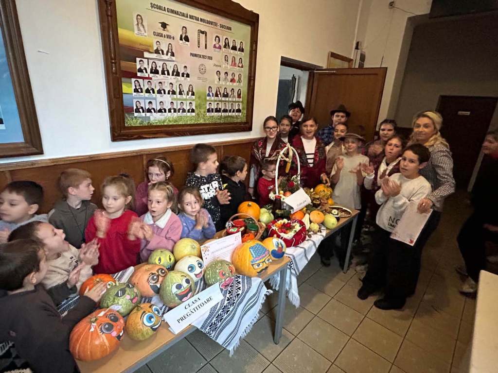 Grup de copii zâmbind și aplaudând lângă o masă plină cu dovleci și fructe decorate, într-o sală de clasă.