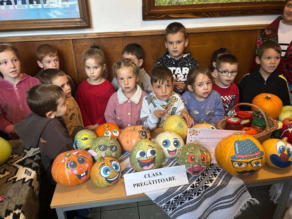 Un grup de copii stând lângă o masă plină cu dovleci pictați și ținând un banner cu textul 'CLASA PREGĂTITOARE'.