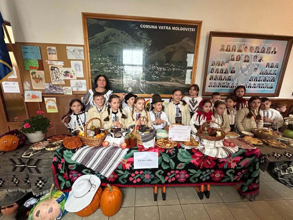 Un grup de copii îmbrăcați în costume tradiționale românești, stând în fața unei mese pline cu mâncăruri tradiționale, la o expoziție culturală în Vatra Moldoviței.