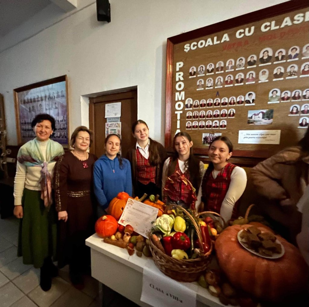 Un grup de femei și fete îmbrăcate în costume populare, stând lângă o masă plină cu legume și fructe, într-un decor interior, cu un tablou de onorare pe perete.