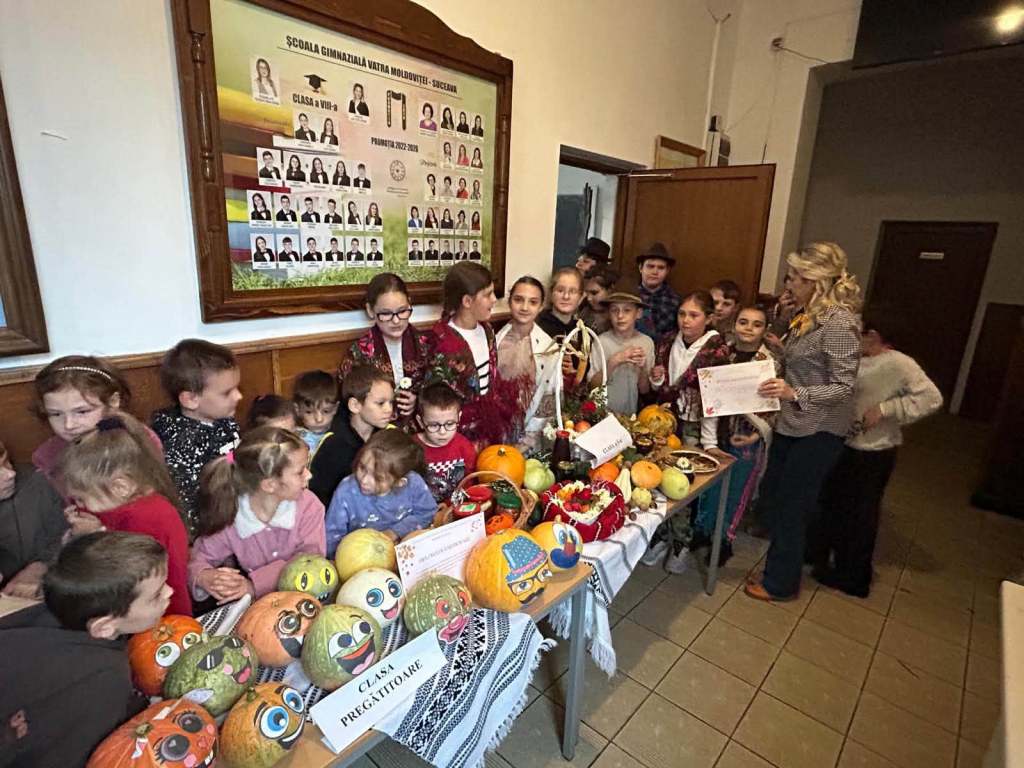 Grup de copii îmbrăcați în costum popular, participând la un eveniment școlar, lângă o masă plină cu dovleci pictați și fructe. În fundal, o fotografie a clasei și un panou informativ.