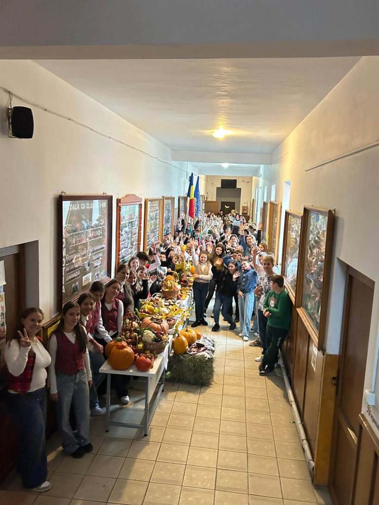 Un grup mare de elevi stând într-un hol, lângă mese pline cu legume și fructe de toamnă, inclusiv dovleci, în timp ce zâmbesc și salută camera.