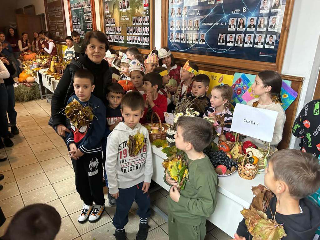 Grup de copii la o activitate școlară, stând lângă mese pline cu păstăi de legume și fructe. O femeie adultă este alături de ei, iar copiii poartă elemente decorative din frunze.