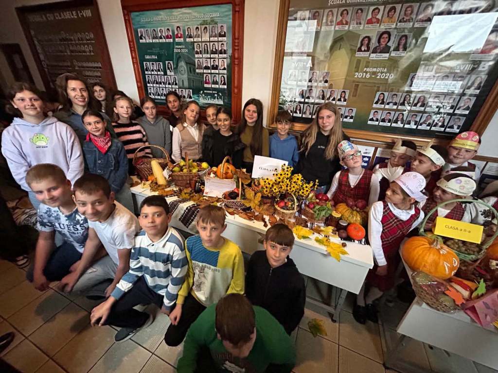 Un grup de copii și adulți pozând împreună, cu mese pline de fructe și legume decorate pentru o activitate școlară, în interiorul unei școli.