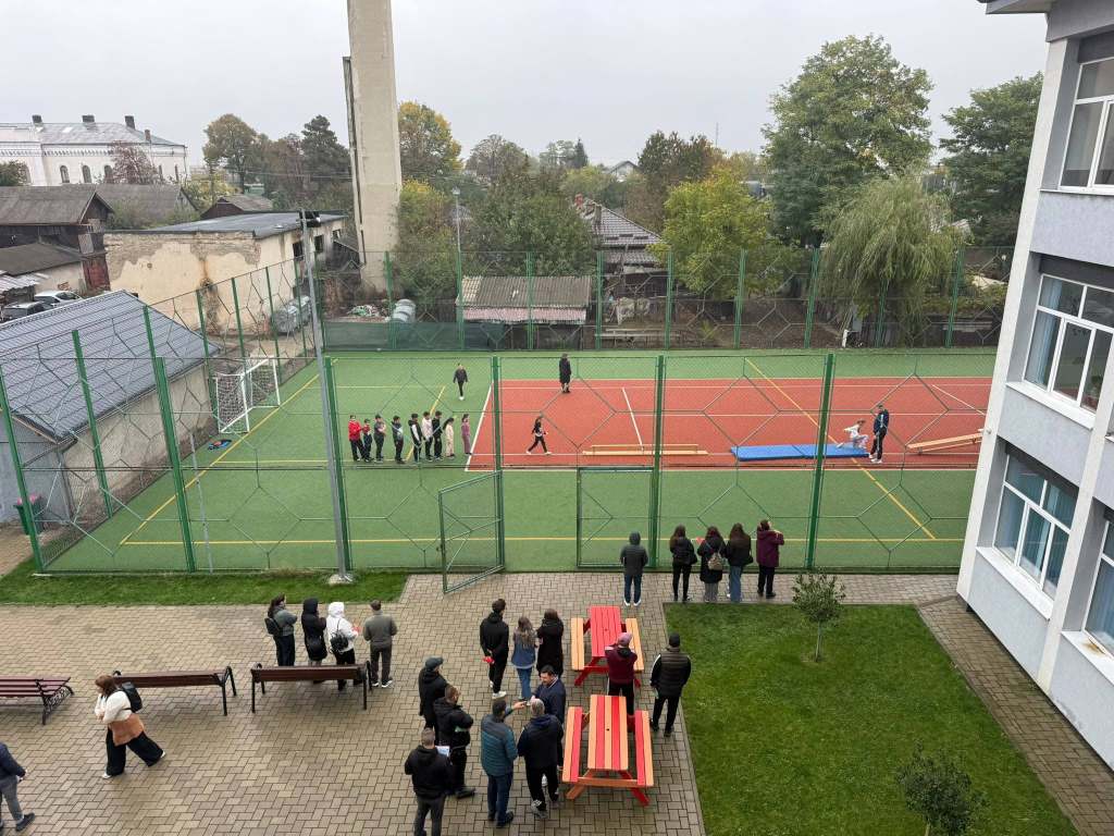 O imagine dintr-o școală, cu un teren de sport acoperit de plasă. Elevi îmbrăcați în uniforme stau în grupuri pe teren, iar câțiva adulți observă de pe margine. În fundal se vede clădirea școlii și copaci.