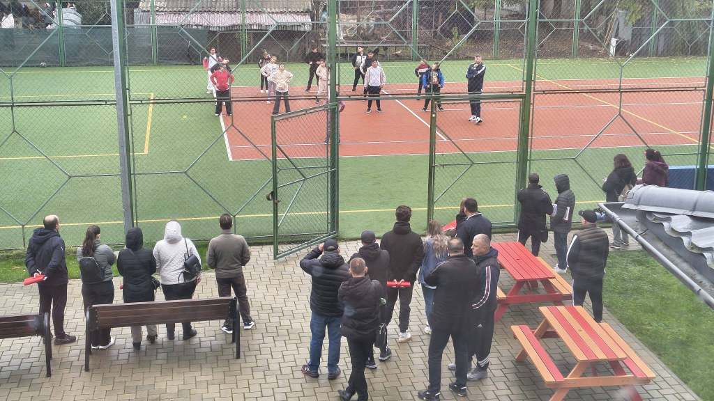 Persoane la un teren de tenis, unele jucând și altele privindu-le, cu bănci și mese în prim-plan.