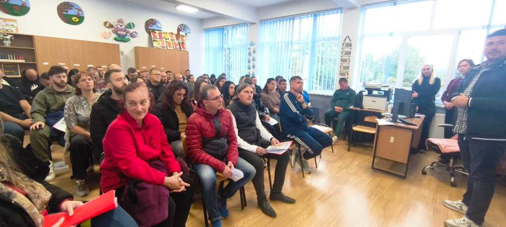 O sală plină de oameni așezati pe scaune, ascultând un vorbitor la o întâlnire sau prezentare. În fundal sunt cărți și un monitor.