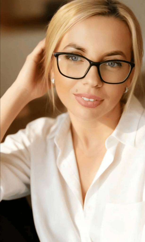Portret al unei femei cu păr blond, purtând o bluză albă și ochelari, zâmbind ușor în lumină naturală