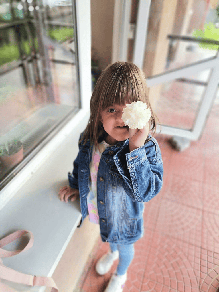 O fetiță cu păr lung și cu breton, îmbrăcată într-o jachetă din denim, ținând o floare albă în fața feței, zâmbind, lângă fereastra unei clădiri.