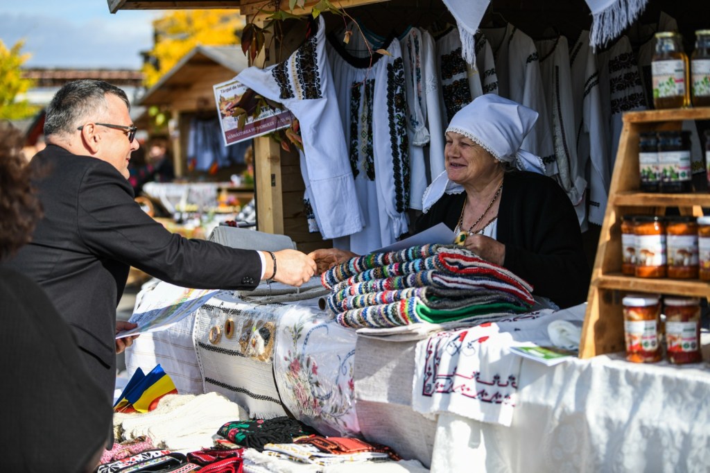 O femeie vinde produse tradiționale la un târg, interacționând cu un bărbat îmbrăcat la costum, în fața unei mese pline cu articole hand-made.