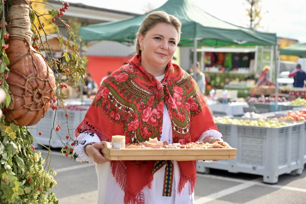 O femeie poartă o ie tradițională și un fular roșu cu motive florale, ținând o tavă cu mâncare. În fundal se află un târg cu tarabe pline de fructe și legume.