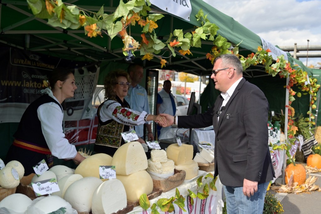 O femeie îmbrăcată în costumul național și un bărbat într-un costum modern se strâng mâna la un stand de brânzeturi, în fața unui cort verde decorat cu frunze.
