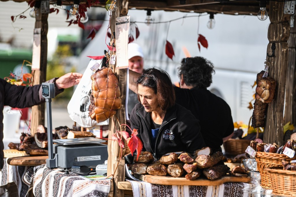 O vânzătoare la un stand de produse din carne, înconjurată de preparate delicioase, cu clienți interacționând.