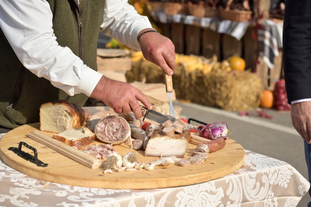 Un bărbat taie delicat carne și pâine pe o masă rotundă din lemn, la un târg de produse alimentare.