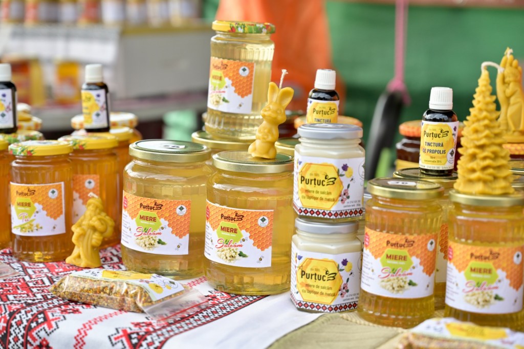 Diverse borcane cu miere și produse apicole, expuse pe o masă, cu etichete colorate. Unii borcane au figurine din ceară de albine, iar în fundal se observă ambalaje variate.