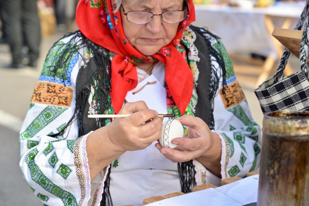 O femeie îmbrăcată în costum popular românesc, cu batic roșu, care decorează un ou cu un penson, concentrată pe detalii.