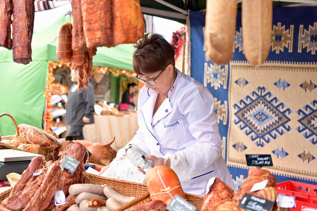 Vânzătoare la un târg de produse tradiționale, pregătind mezeluri într-un stand decorat cu un covor tradițional.