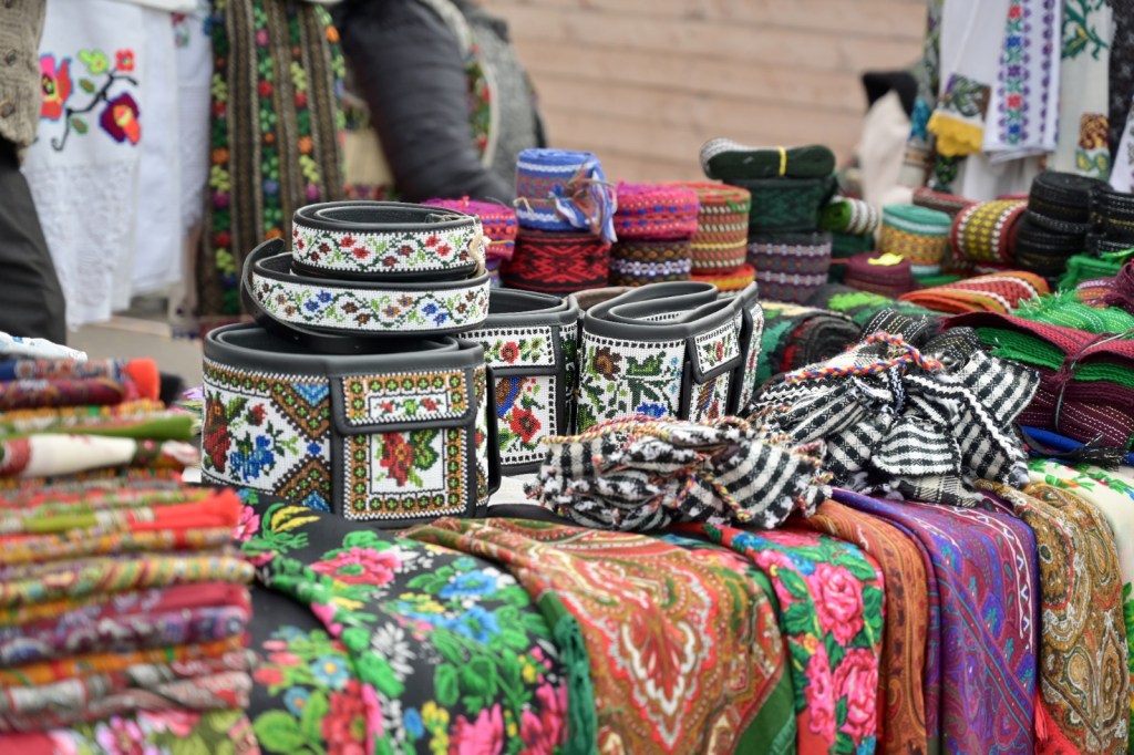 Târg cu articole tradiționale, inclusiv centuri și materiale textile colorate.