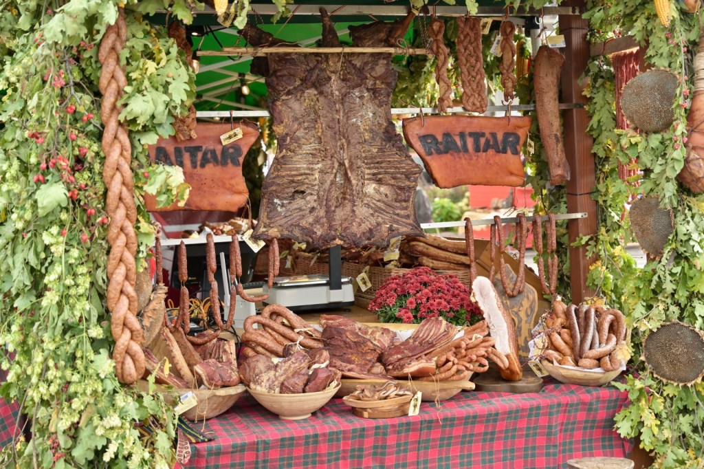 Stand de produse din carne cu diverse sortimente expuse, inclusiv mezeluri și carne afumată, împodobit cu plante și flori.