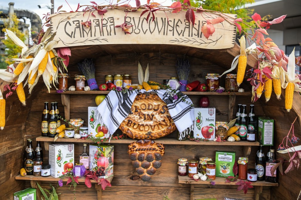 Un stand decorat cu produse tradiționale românești, inclusiv borcane cu dulcețuri, sucuri și băuturi, aromatizat cu flori și porumb.