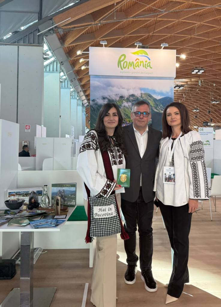 Trei persoane îmbrăcate în costume populare românești, pozând în fața unui stand despre România la un eveniment. Bannerul de sus promovează România ca destinație turistică.