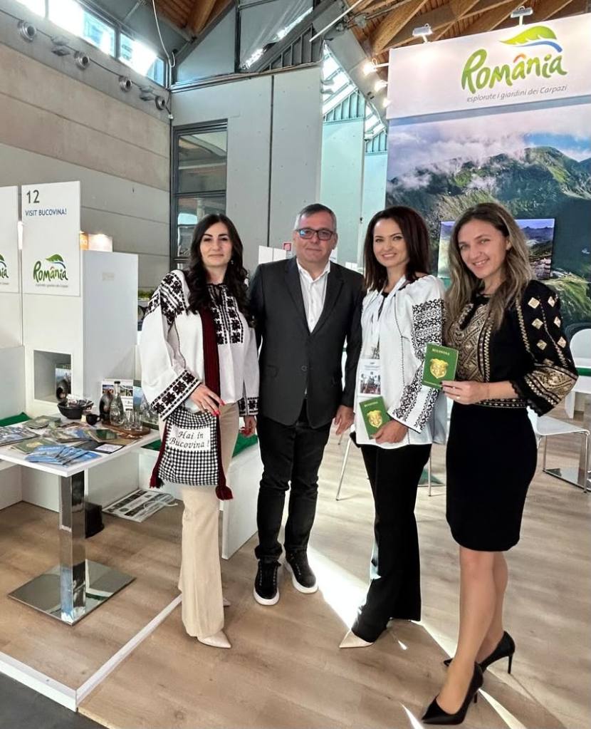 Grup de patru persoane îmbrăcate în costume tradiționale românești la un stand turistic dedicat României, cu pliante și informații despre Bucovina în fundal.