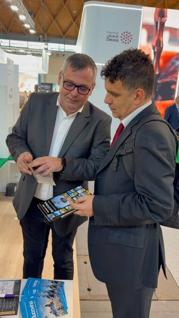 Doi bărbați discută la un stand, examinând un catalog și brosuri de informații.