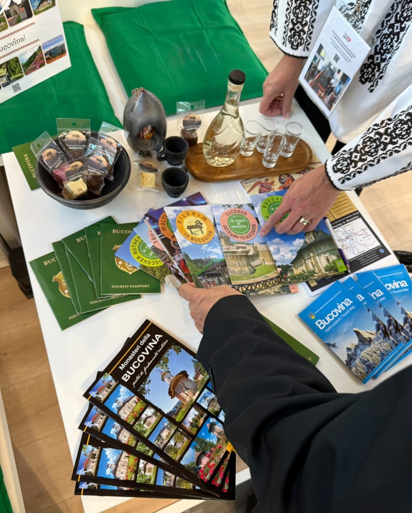 Mână arătând spre pliante și produse turistice din Bucovina, cu sticlă de băutură și pahare pe o masă.