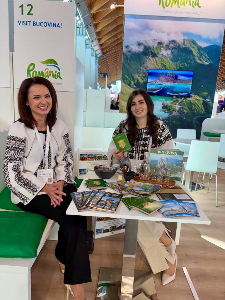 Două femei stând la o masă cu materiale promoționale despre Bucovina, într-un stand reprezentativ pentru România, cu un panou care încurajează vizitarea regiunii.