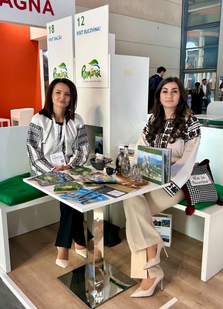 Două femei stând la o masă într-un stand de expoziție dedicat turismului în România, cu pliante și suveniruri pe masă.