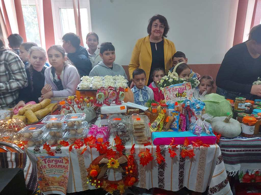 O femeie și copii stând lângă o masă plină cu alimente de sezon, la un târg de toamnă. Masa este decorată cu diverse produse, cum ar fi fructe, gemuri și dulciuri.