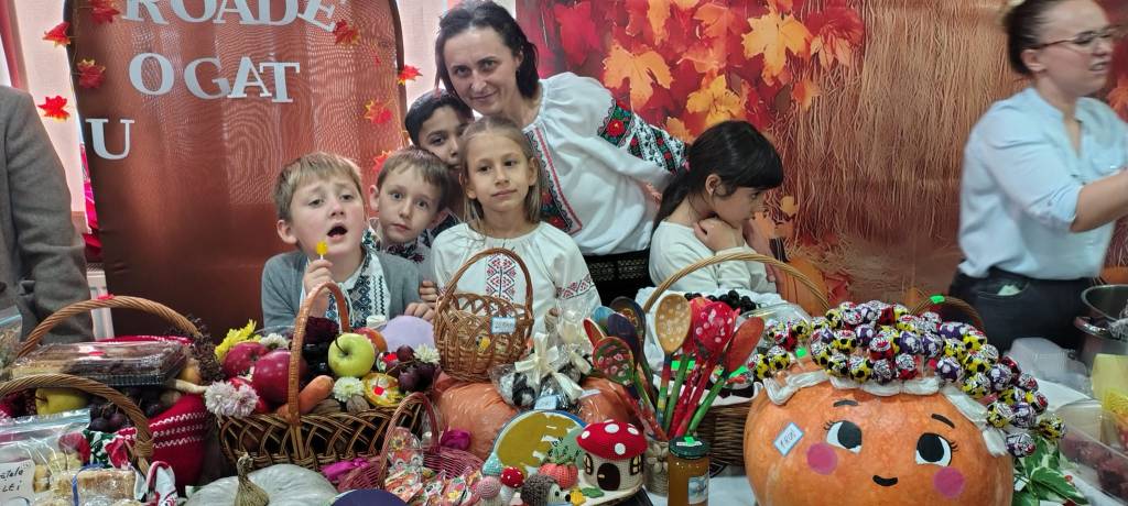 Un grup de copii și adulți lângă un stand plin de produse tradiționale, cum ar fi fructe, legume, și dulciuri, cu fundal de toamnă.