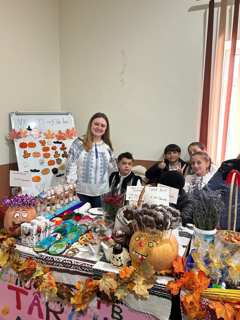 O femeie și patru copii stau lângă un stand plin cu produse artizanale, inclusiv decorațiuni de Halloween, dulciuri și flori uscate, în cadrul unei expoziții.