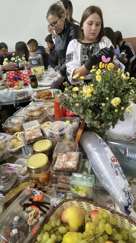 O masă plină de delicii culinare tradiționale, cu diverse preparate, dulcețuri și fructe, iar în fundal persoane implicate în activități comunitare.